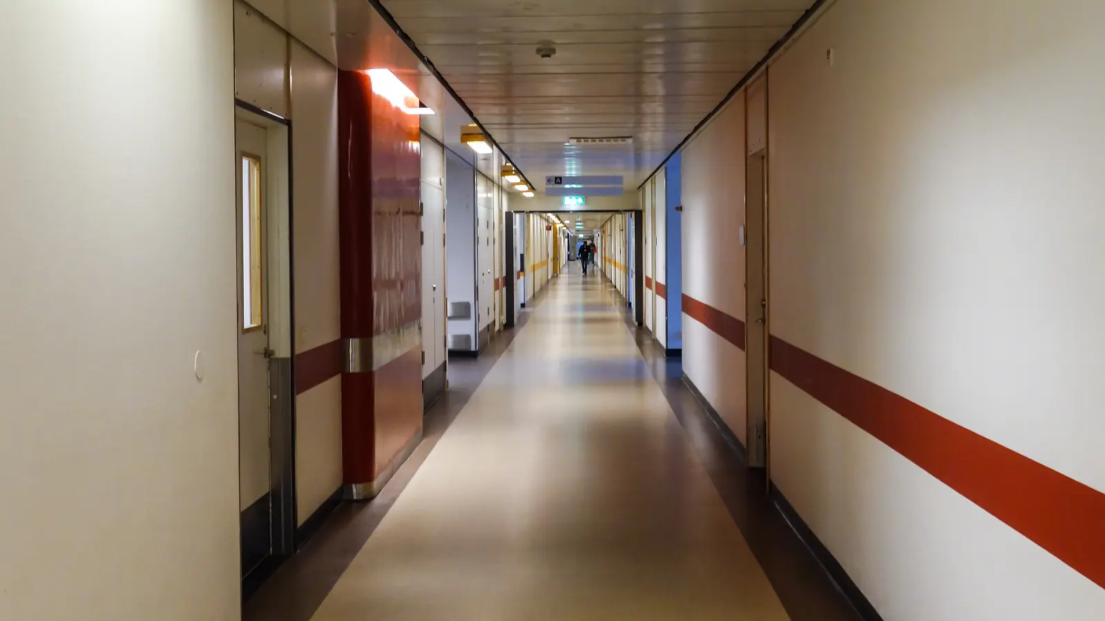 A long hospital corridor with a red stripe along the wall, receding towards a lit doorway