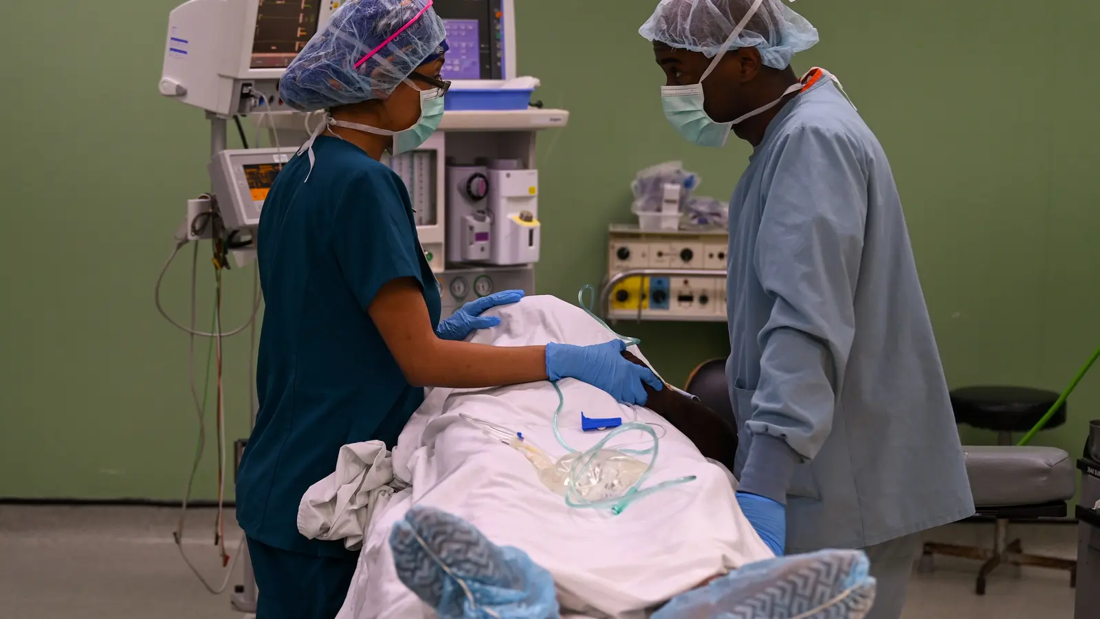 Two clinicians in scrubs and surgical caps attending to a draped patient in an operating room