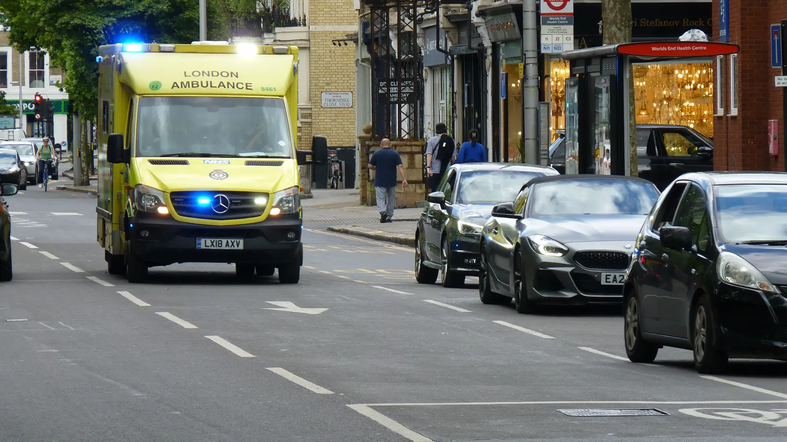 A London Ambulance Service vehicle on blue lights passing a health centre on a city street