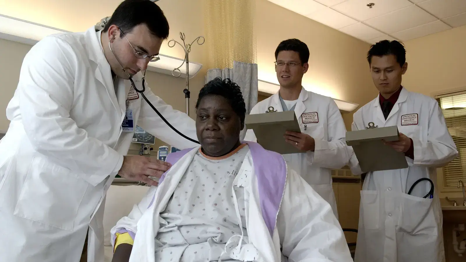 A doctor examining a seated patient with a stethoscope while two colleagues observe and take notes