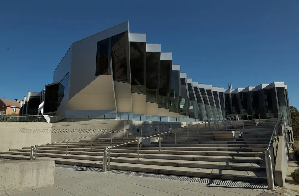 Exterior of the John Curtin School of Medical Research (ANU's medical school building), Canberra, Australia — angular metallic-and-glass facade with grand staircase under a clear blue sky.
