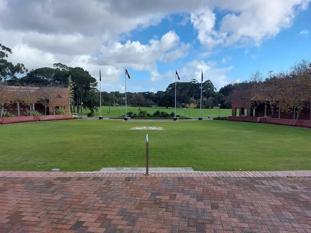 The Forum central lawn on Curtin University's Bentley (Perth) campus, viewed from Whitlam Walk — flags, green lawn, red-brick colonnades under a blue sky.