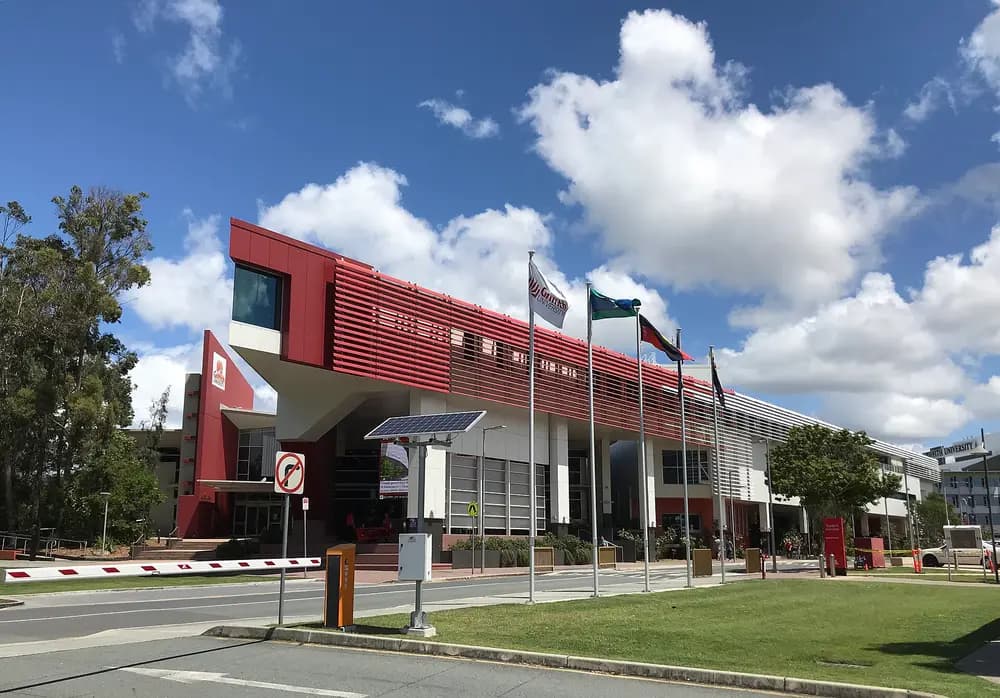 Daytime exterior of the striking red-and-white Griffith University building (Library/Student Services) on the Gold Coast campus in Queensland, Australia — the campus hosting the School of Dentistry and Oral Health — under a blue sky with Griffith University flags.