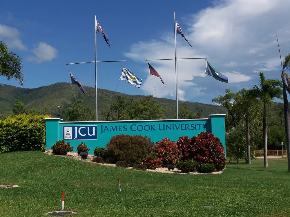 James Cook University campus entrance monument with tropical palms, flags and the Mount Stuart bushland backdrop under a bright blue sky (Townsville, the JCU flagship campus).