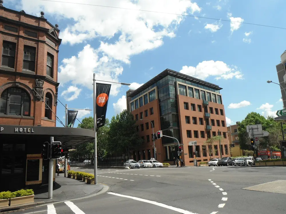 Daytime exterior street view of the University of Notre Dame Australia building at the Oxford Street, Darlinghurst campus in Sydney (home of the Sydney School of Medicine), a modern brick-and-glass building under a bright blue cloudy sky.