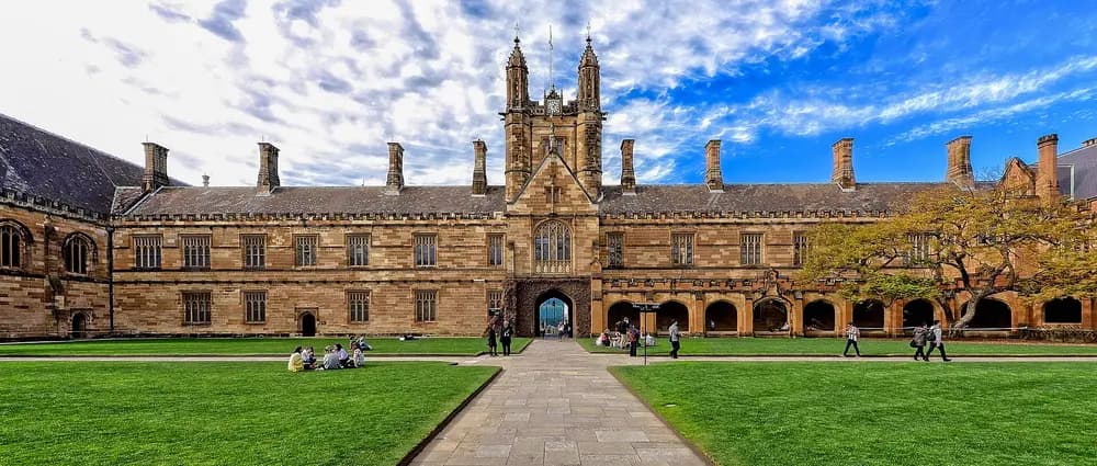 The iconic sandstone Gothic Revival Main Quadrangle and clock tower of the University of Sydney (the host university of its School of Dentistry), Sydney, Australia — wide symmetrical daytime exterior with green lawn and dramatic sky.